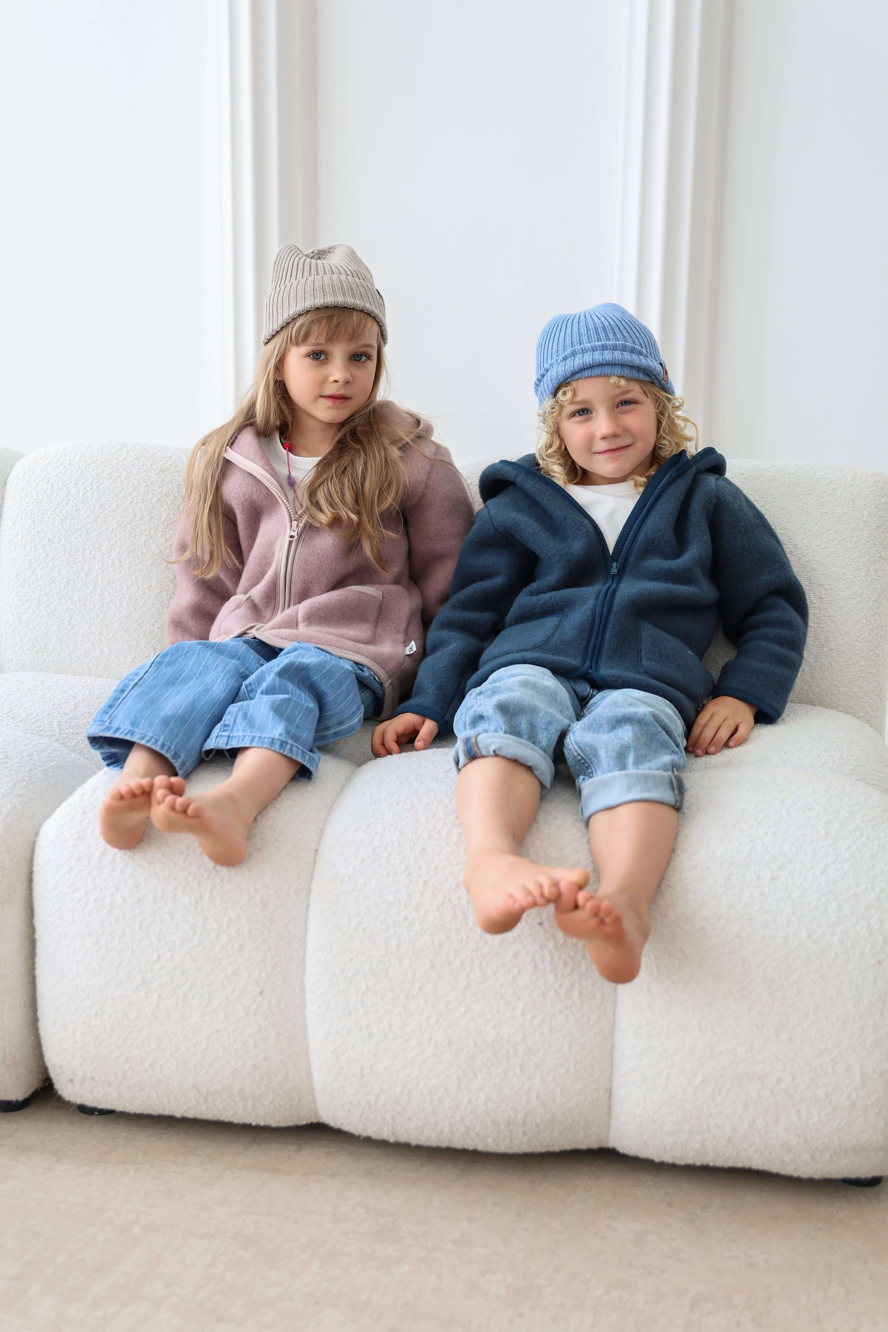 Two children sitting on a white couch wearing matching outfits with hats