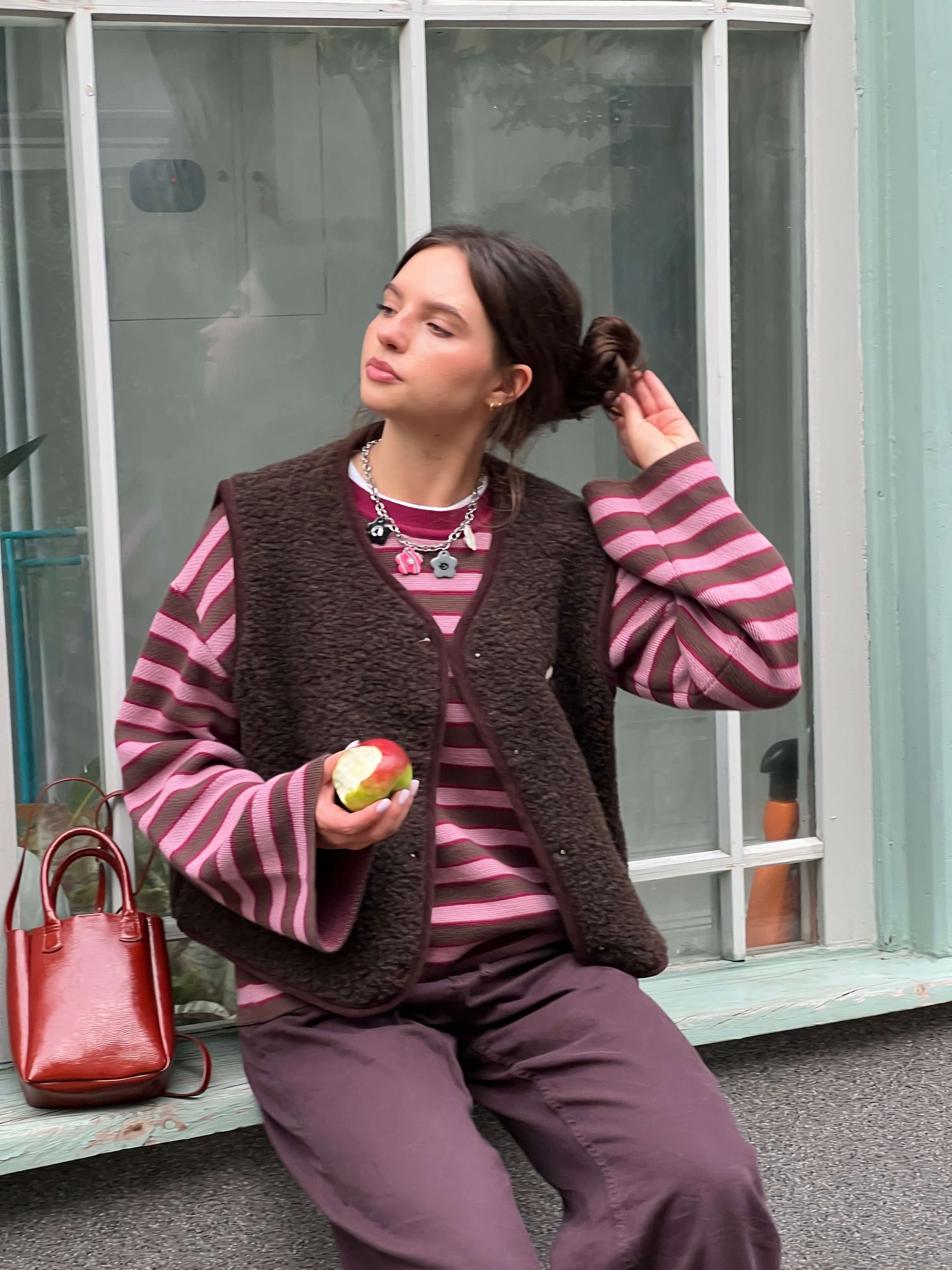 Woman in a striped sweater and vest sitting on a bench holding an apple