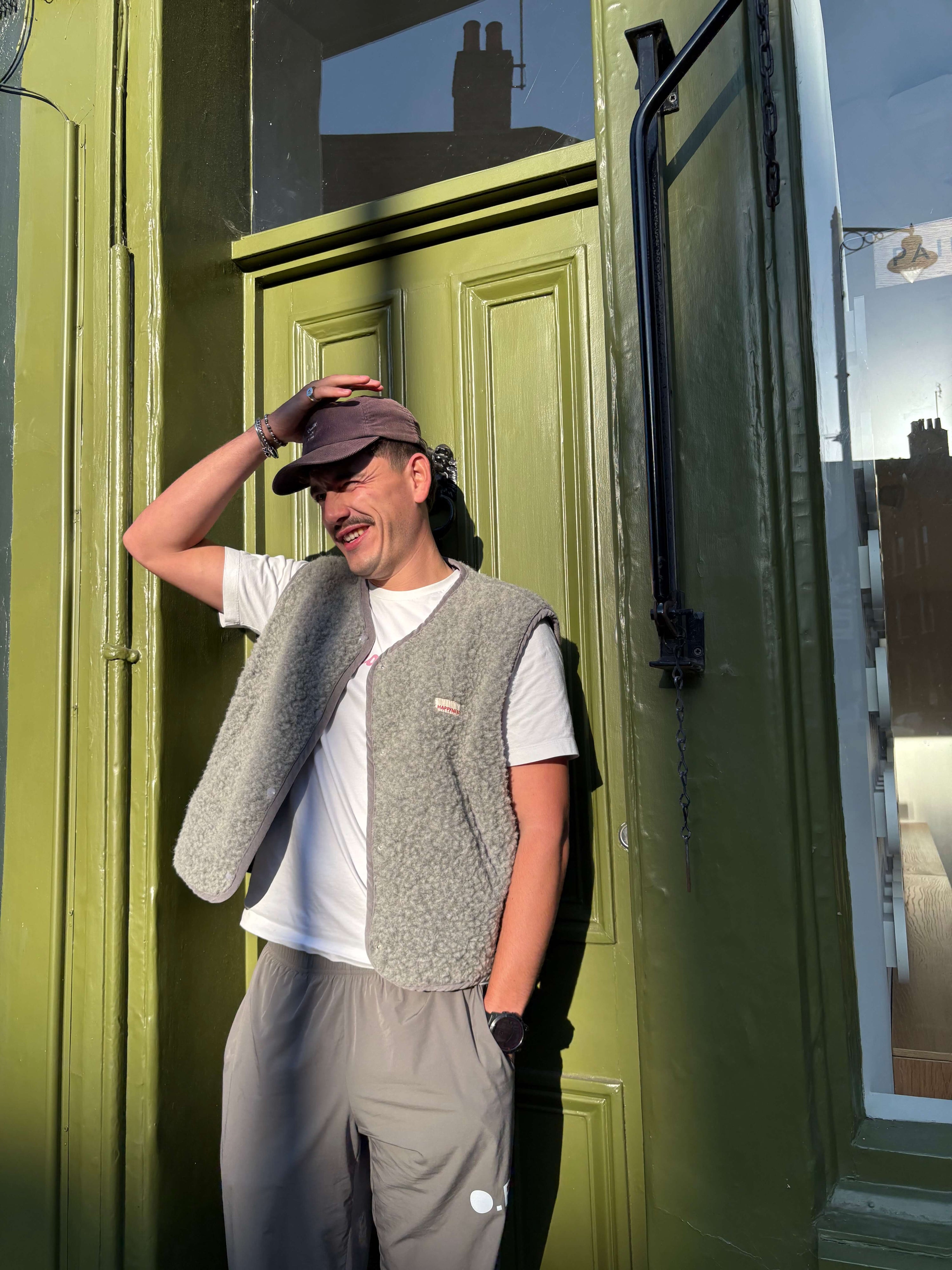 Man standing in front of a green door wearing a gray vest and cap