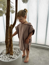 Child in a brown towel standing next to a potted plant indoors