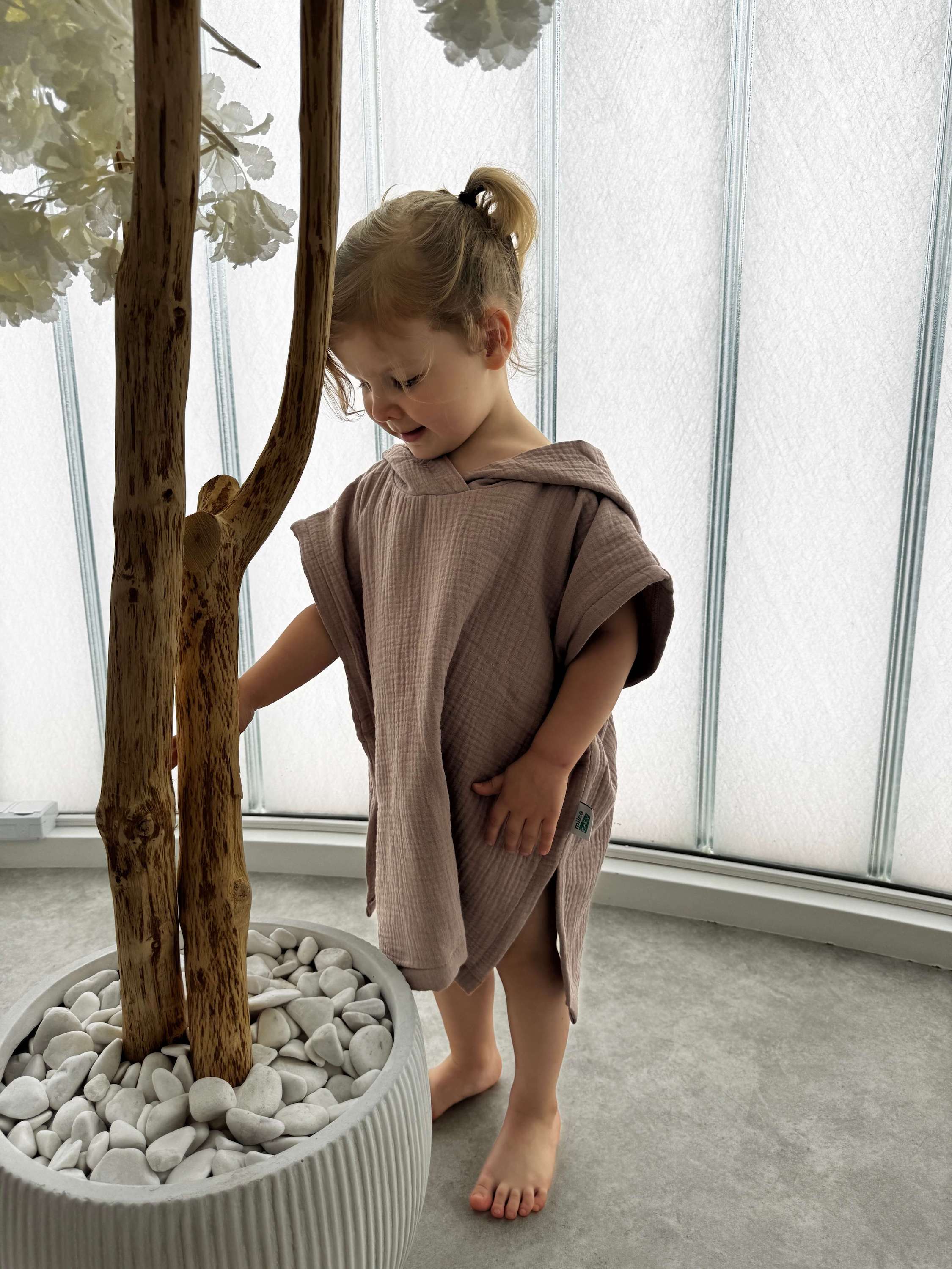 Child in a brown towel standing next to a potted plant indoors