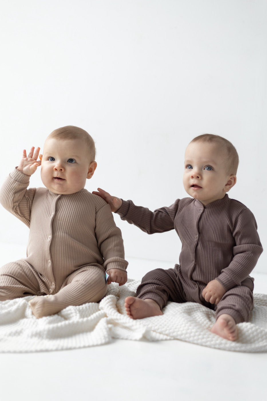 Two babies sitting on the white blanket wearing ribbed rompers, one with feet in color beige, one without feet in color chocolate