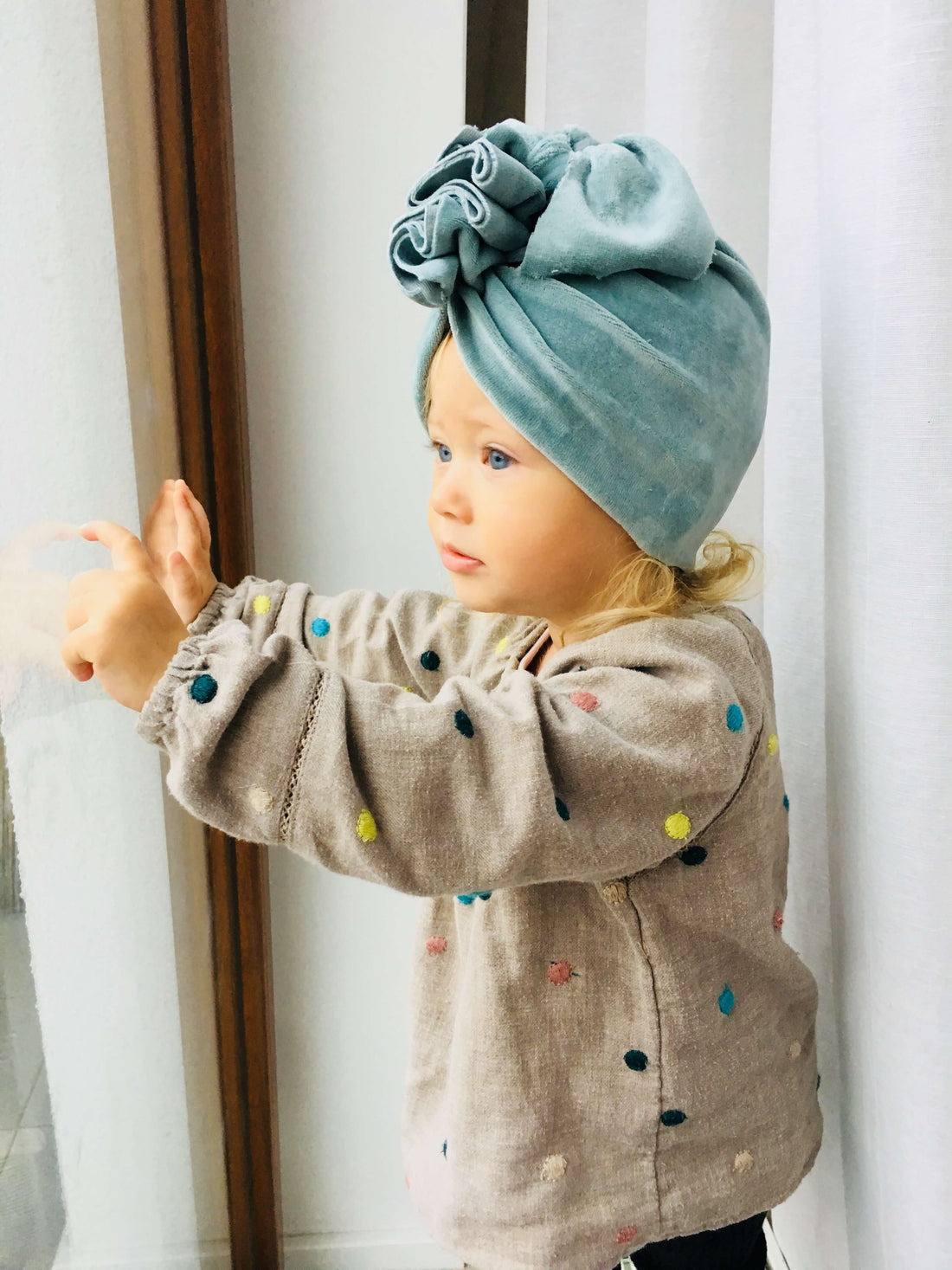 A girl wearing a velvet dusty mint turban and blouse with colorful polka dots looking out of the window