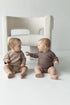 zwei Babys sitzen auf dem Boden vor dem Sessel. Sie habn beige und braue Wickelbody an.