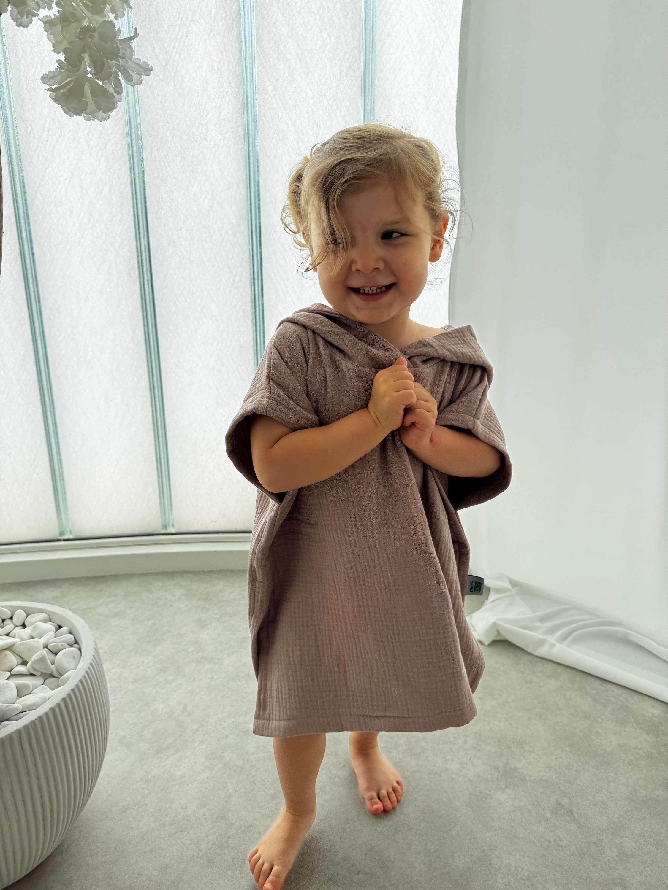 Child wearing a brown poncho in a bright room with large windows.
