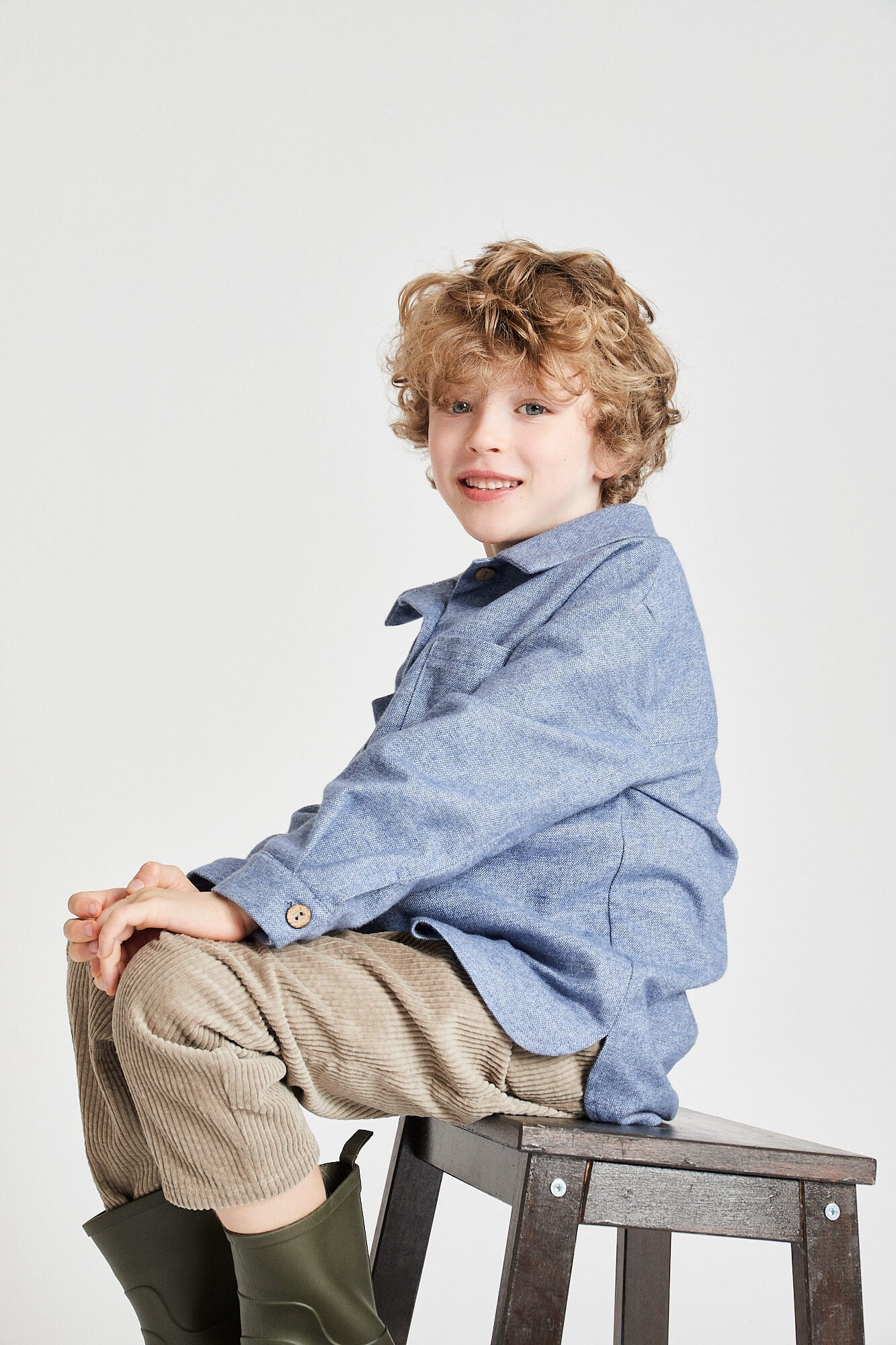 Young boy sitting on a stool wearing a long-sleeved blue shirt and beige pants