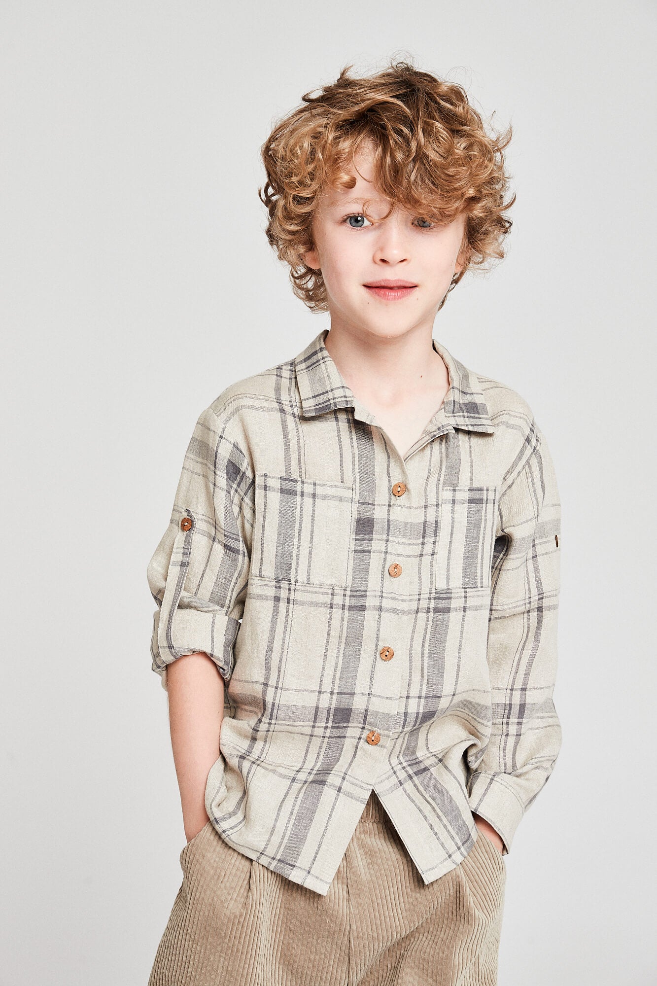 Young boy wearing a beige checkered classic linen shirt with a round neckline and button-down front, paired with brown trousers