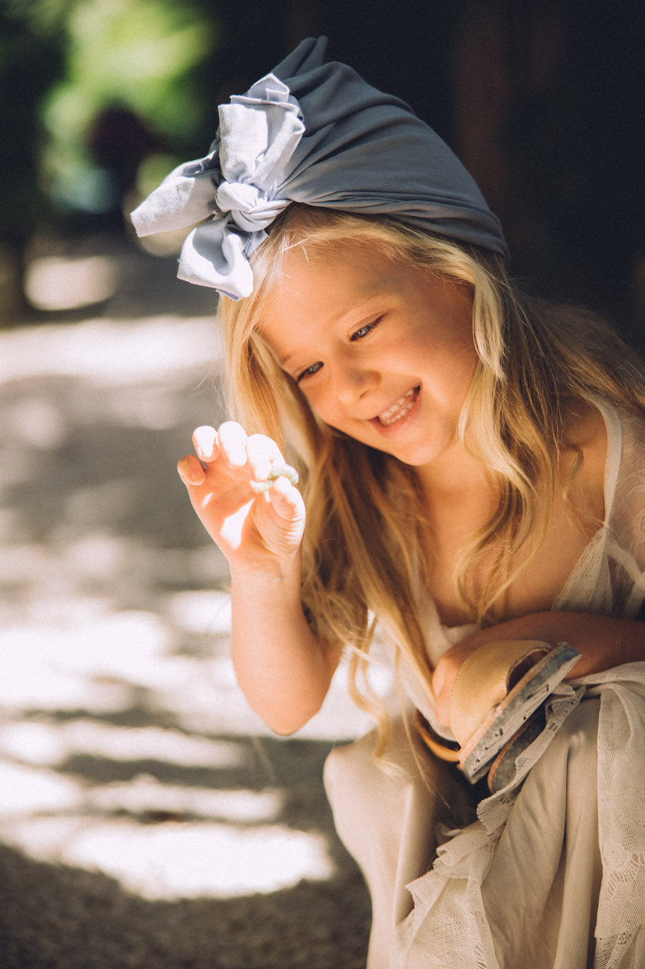 A smiling girl wearing a light blue turban holding her shoes in her hand and looking at a stone in her hand.