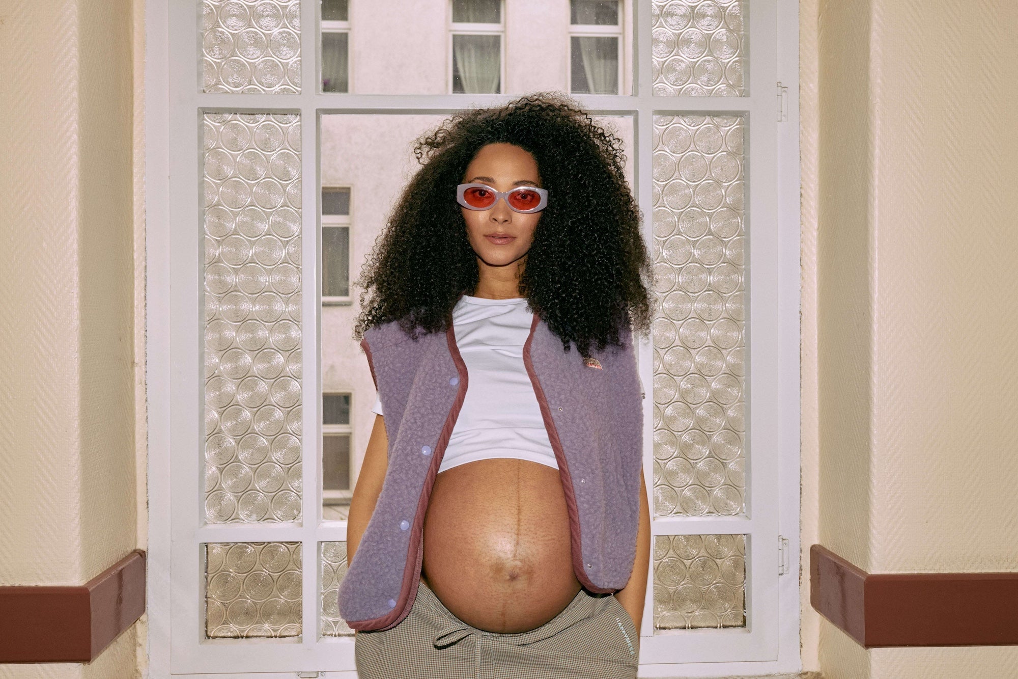 Pregnant woman wearing sunglasses and a purple vest standing in front of a window.