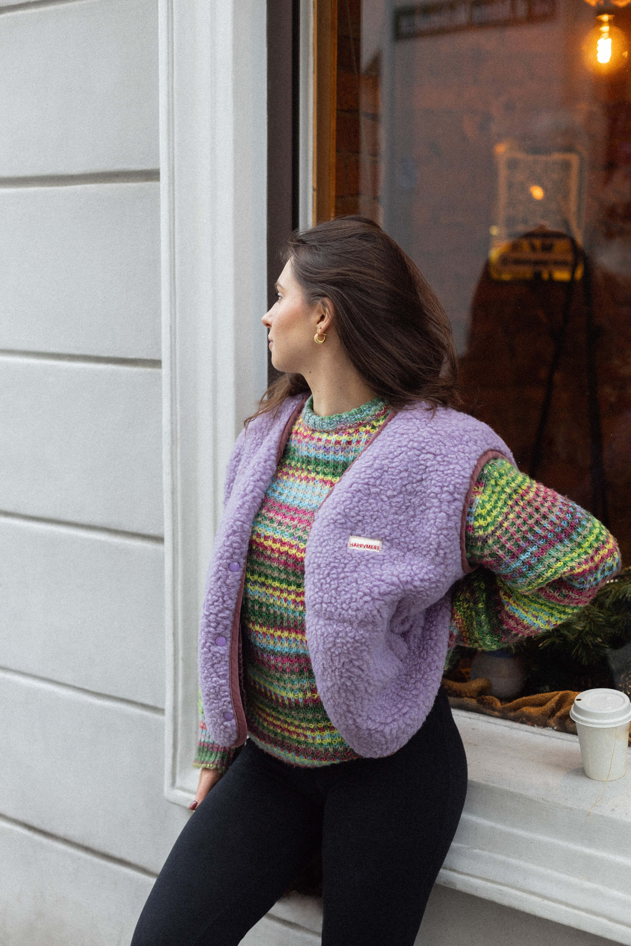 Woman wearing a colorful sweater and purple jacket leaning against a window.