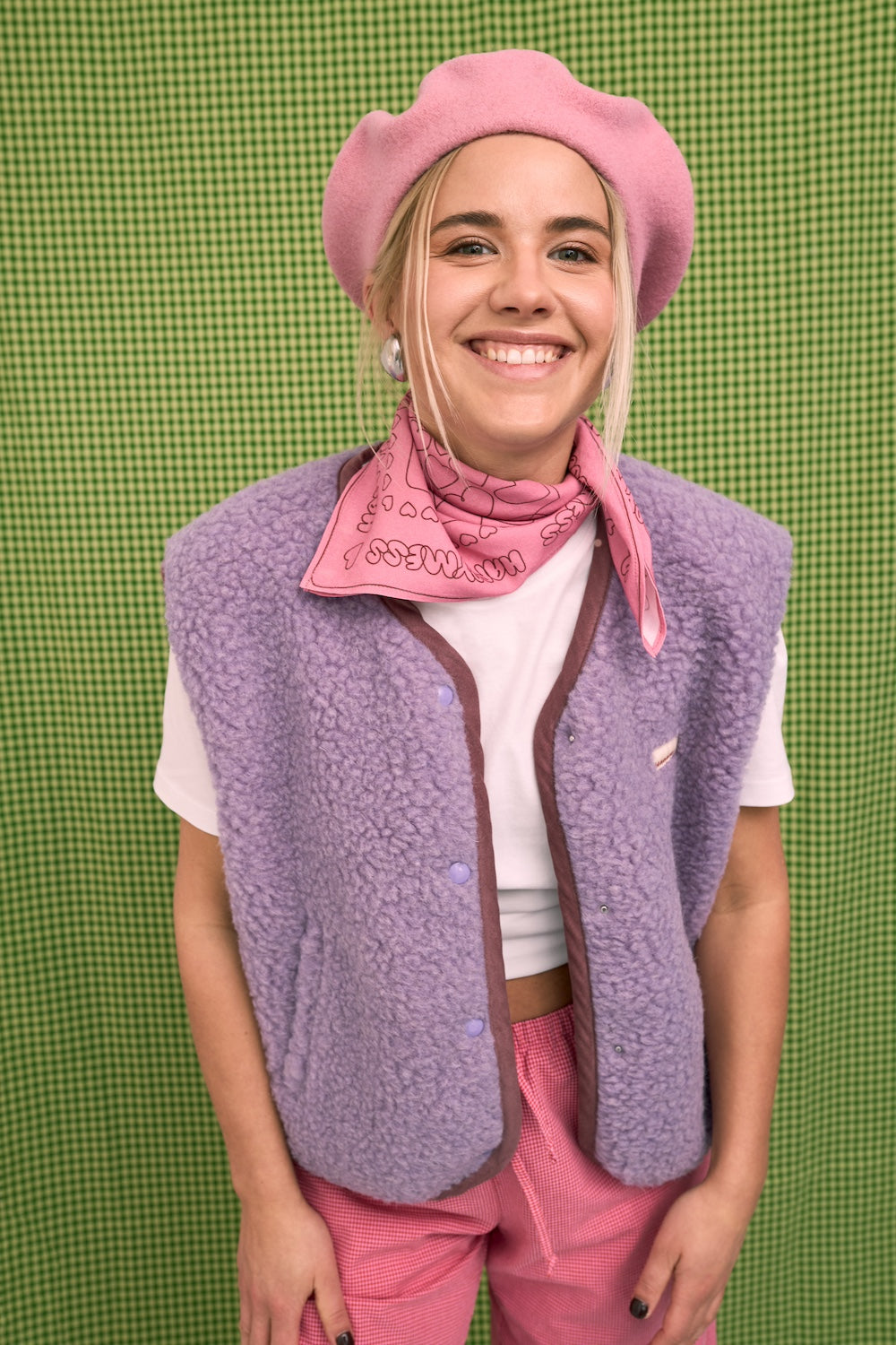 Person wearing a pink beret, scarf, and vest against a green checkered background