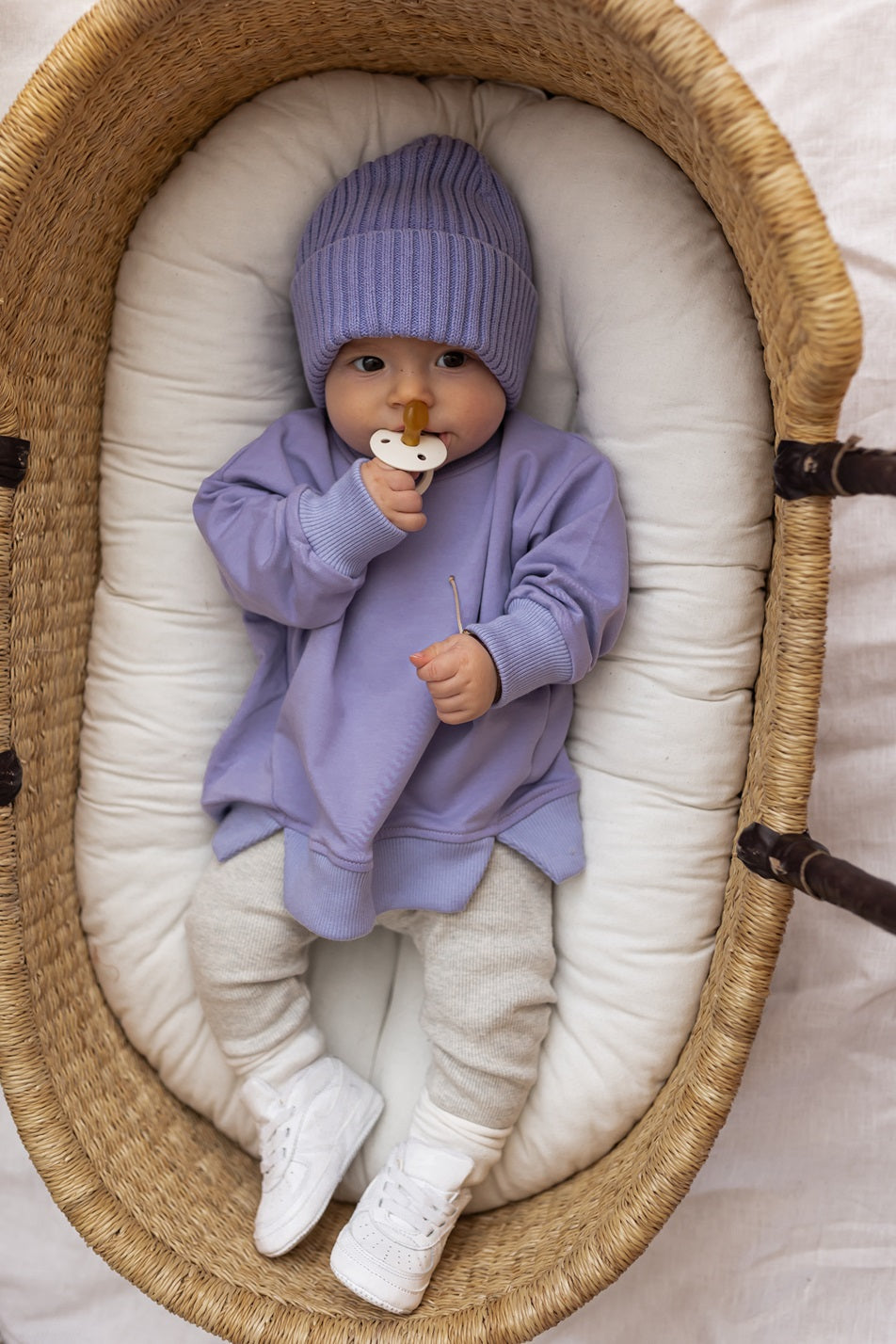 Baby wearing a lilac sweatshirt and oat leggings lying in a wicker basket