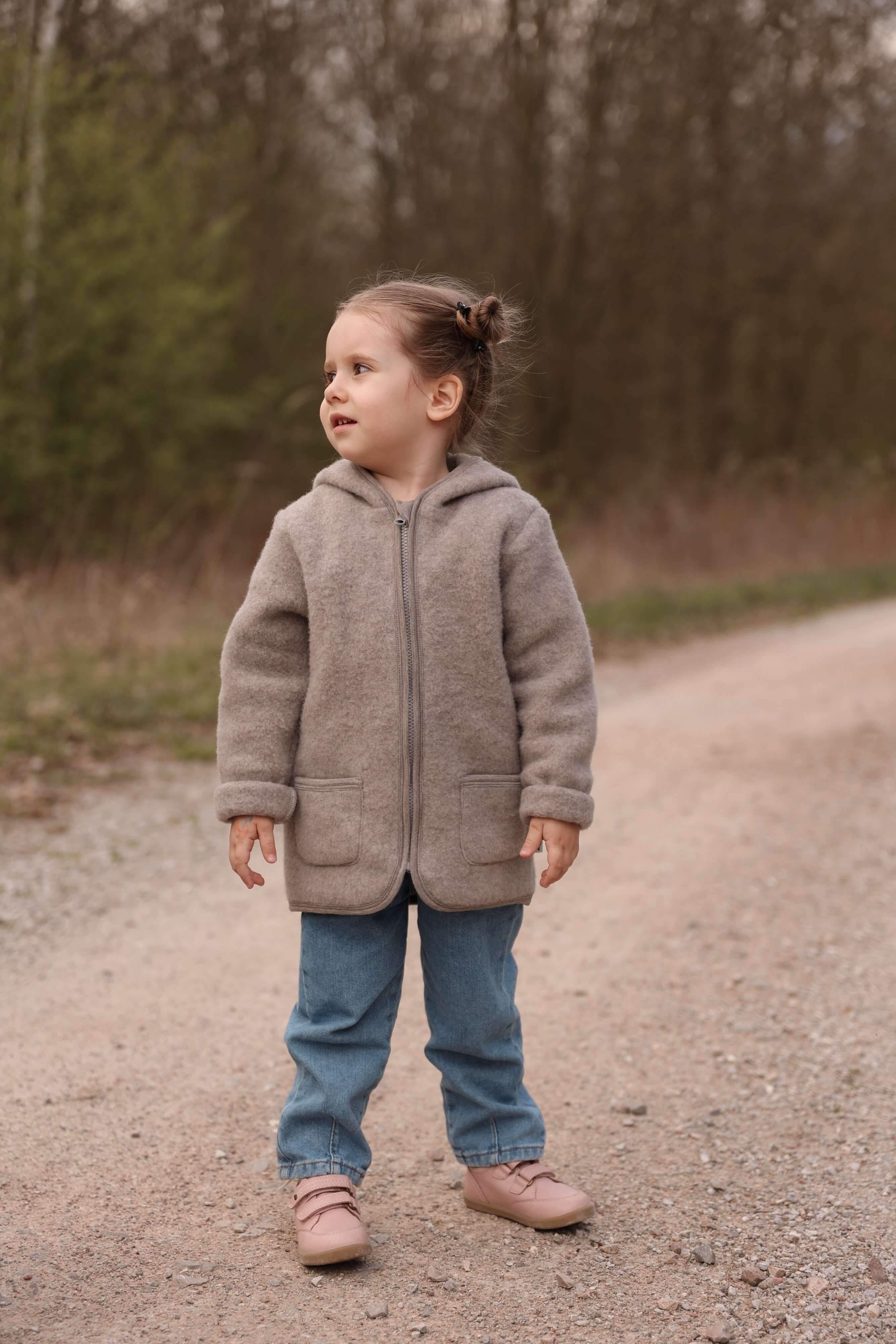 Child wearing a brown jacket standing on a dirt path with trees in the background