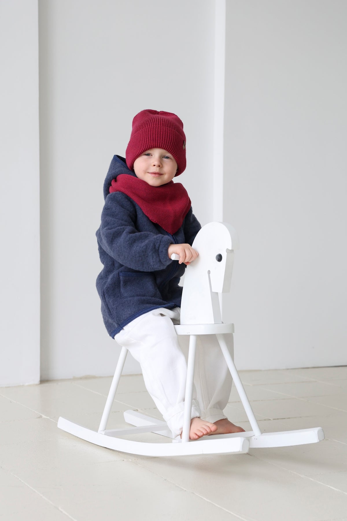 A young child wearing a navy blue hooded sweatshirt, a red knit beanie, and a red neck warmer sits barefoot on a white rocking horse in a bright, minimalist room.