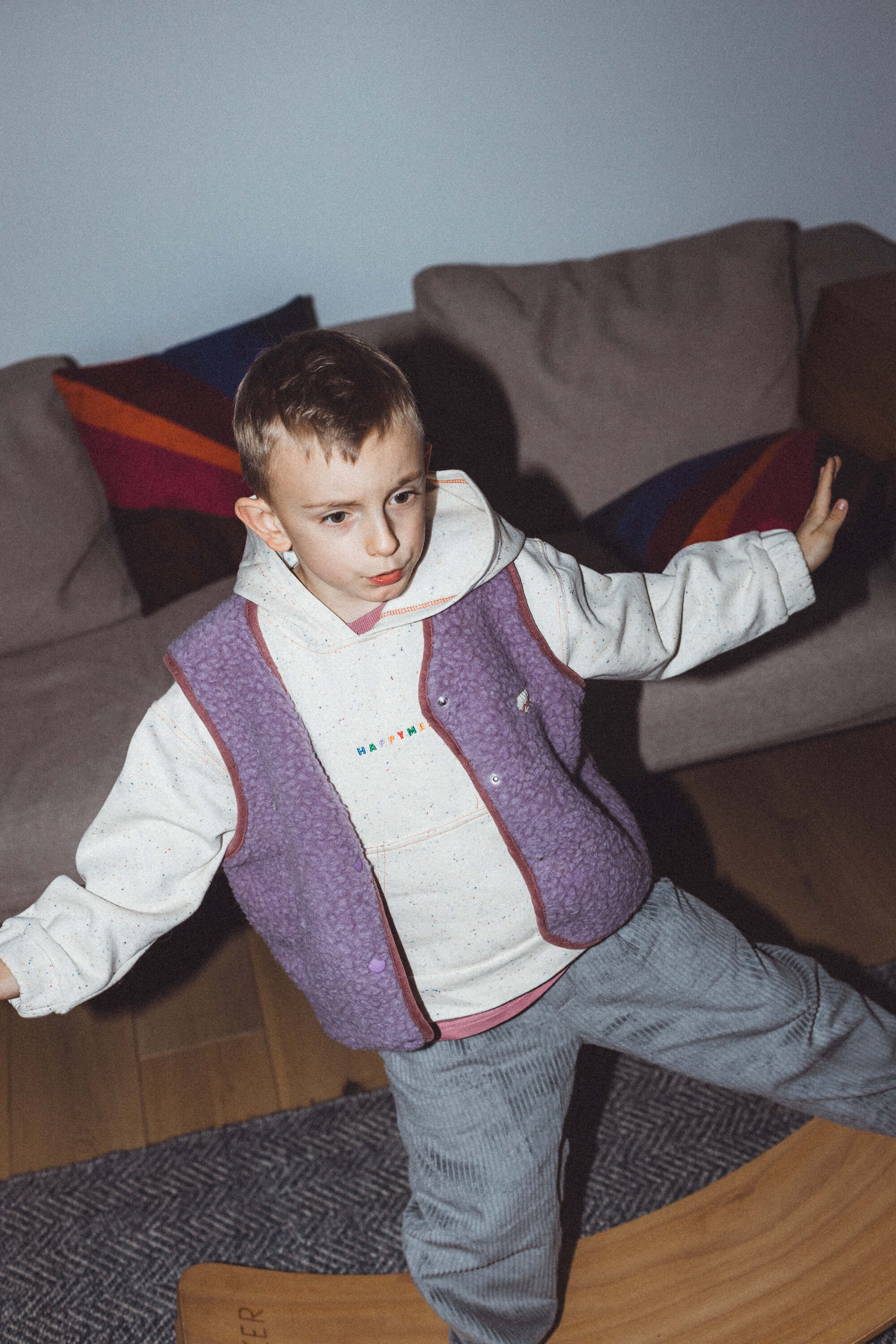 A child standing, wearing a long sleeveless purple merino wool vest with a round neckline, over a white sweatshirt, paired with gray pants.