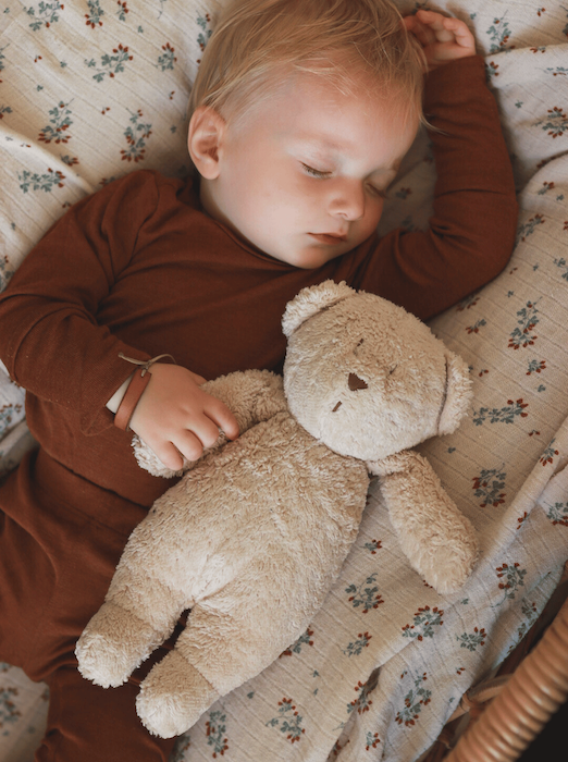 Ein schlafendes Baby in braunem Body und Leggins aus Merinowolle und Seide. Das Baby haltet ein Bär.