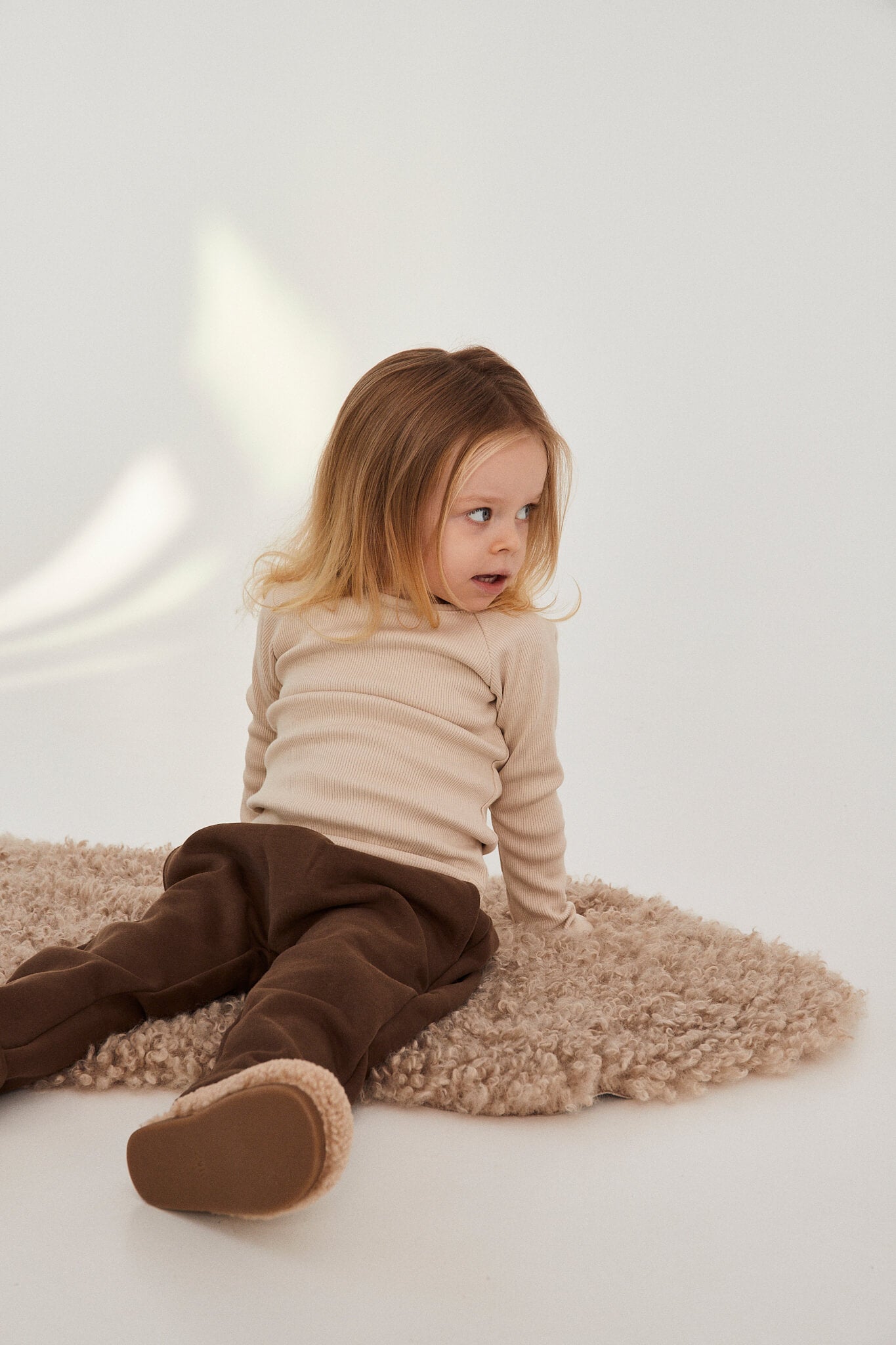 Child sitting on a textured rug wearing a beige sweater and brown pants