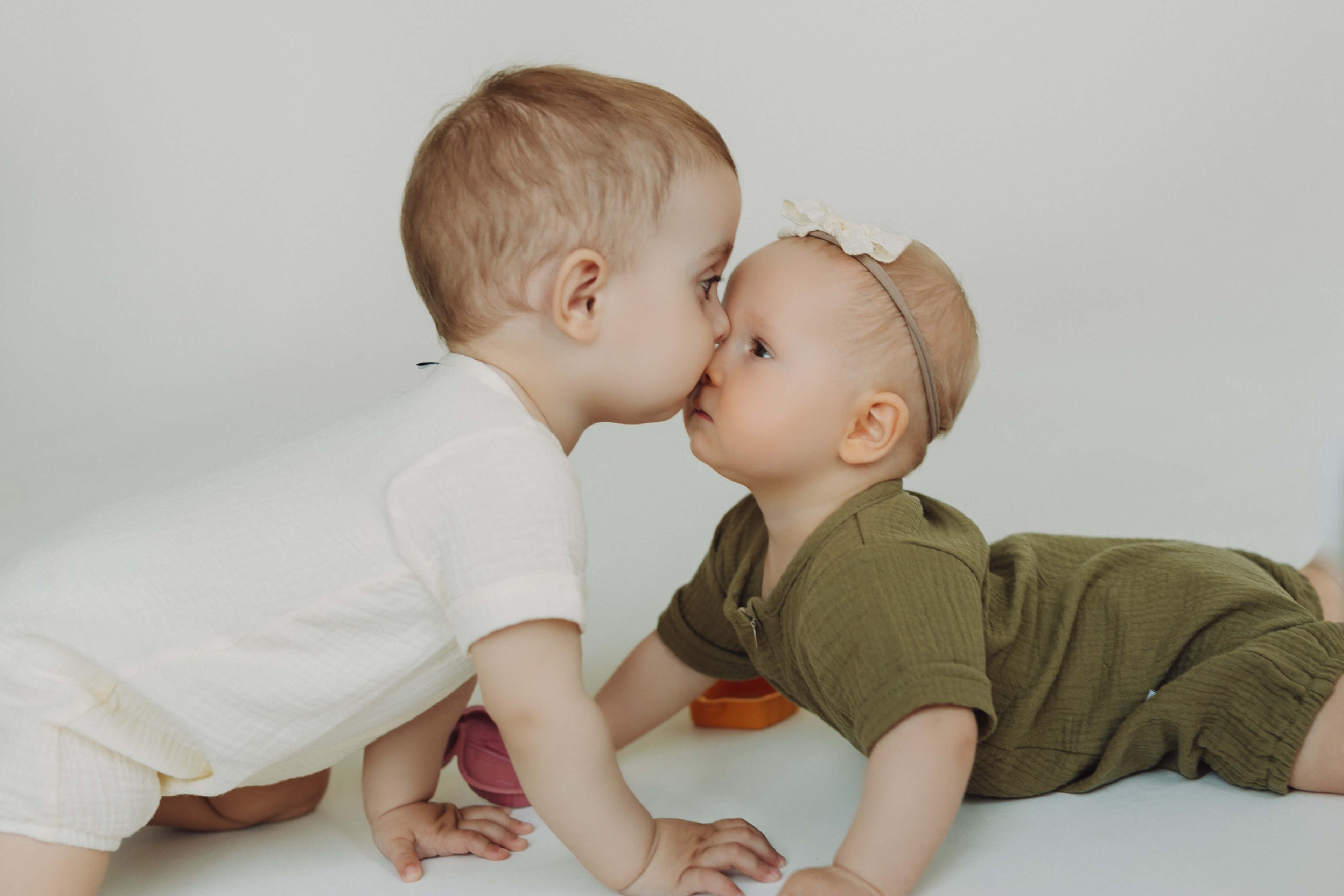 Two babies in white and green outfits lying on a light surface, facing each other.