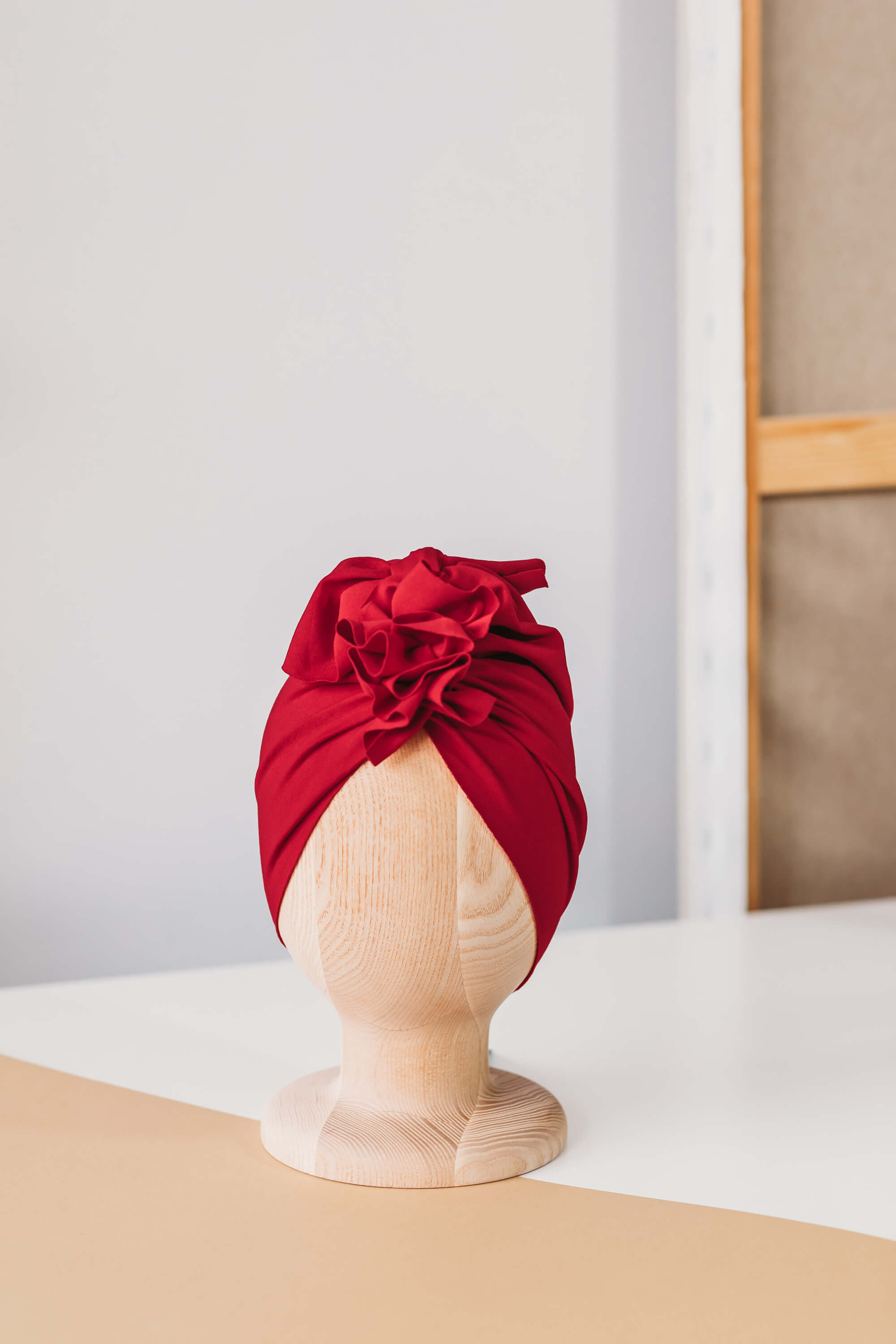 A red turban for children on a wooden mannequin on a white and beige counter with wooden white frame in the background