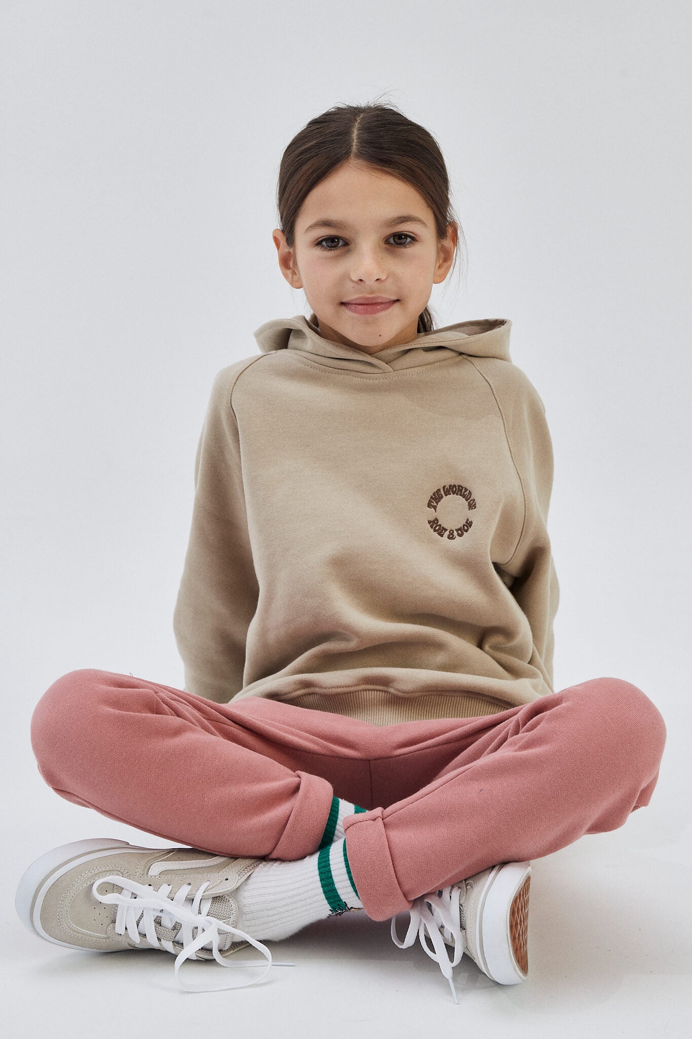 A child sitting cross-legged wearing pink organic cotton sweatpants with an elastic waistband and white sneakers