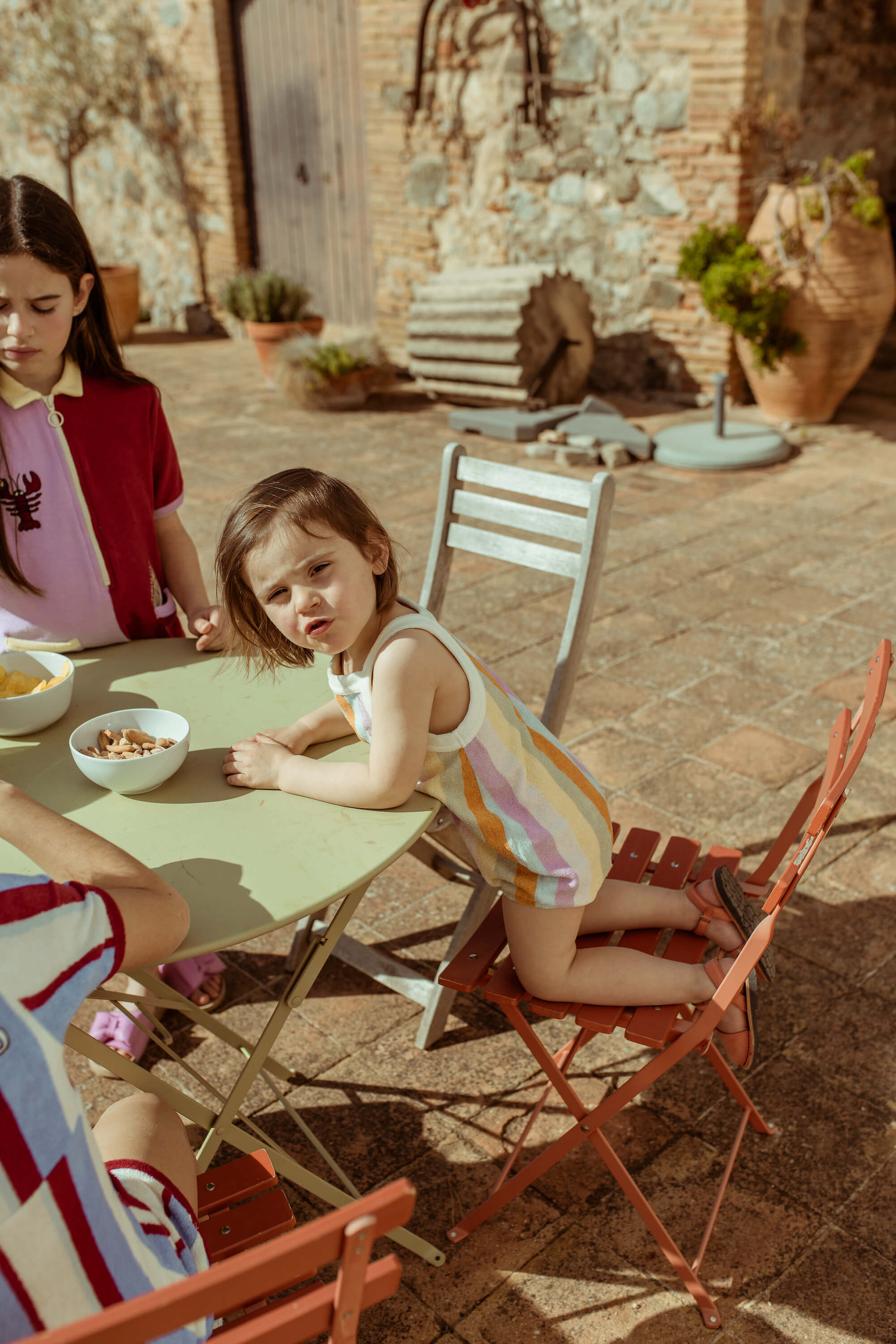 A child sitting at an outdoor table wearing a pink and white striped romper with a palm tree detail, accompanied by an older girl.