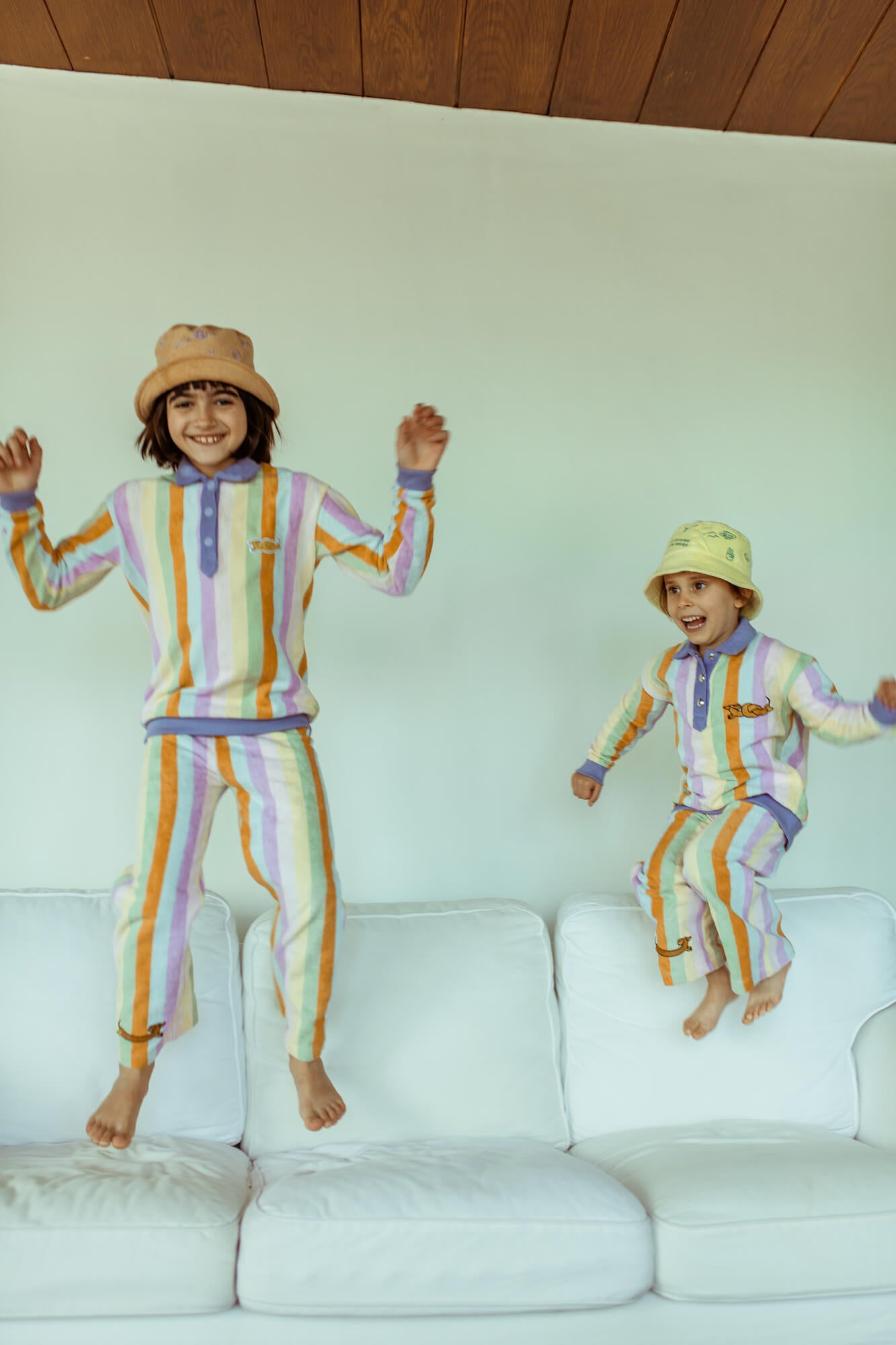 Two children in matching colorful outfits jumping on a white couch.