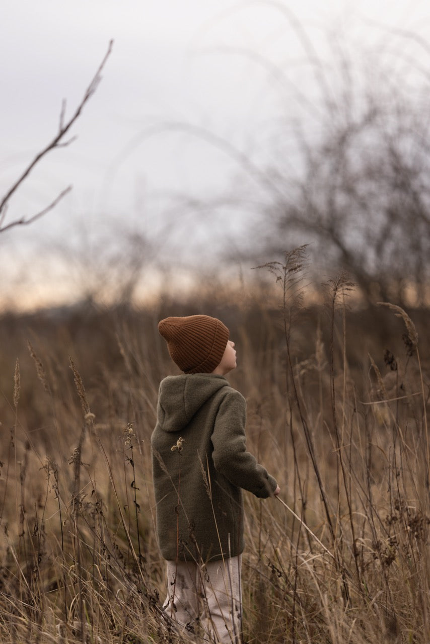 Child in a green jacket and brown beanie standing in tall, dry grass