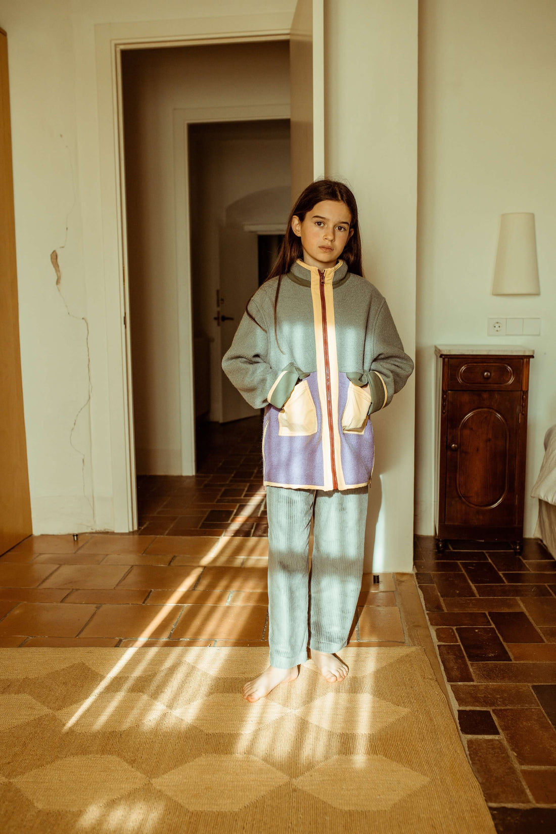 Young girl standing in a sunlit room wearing a patterned outfit.