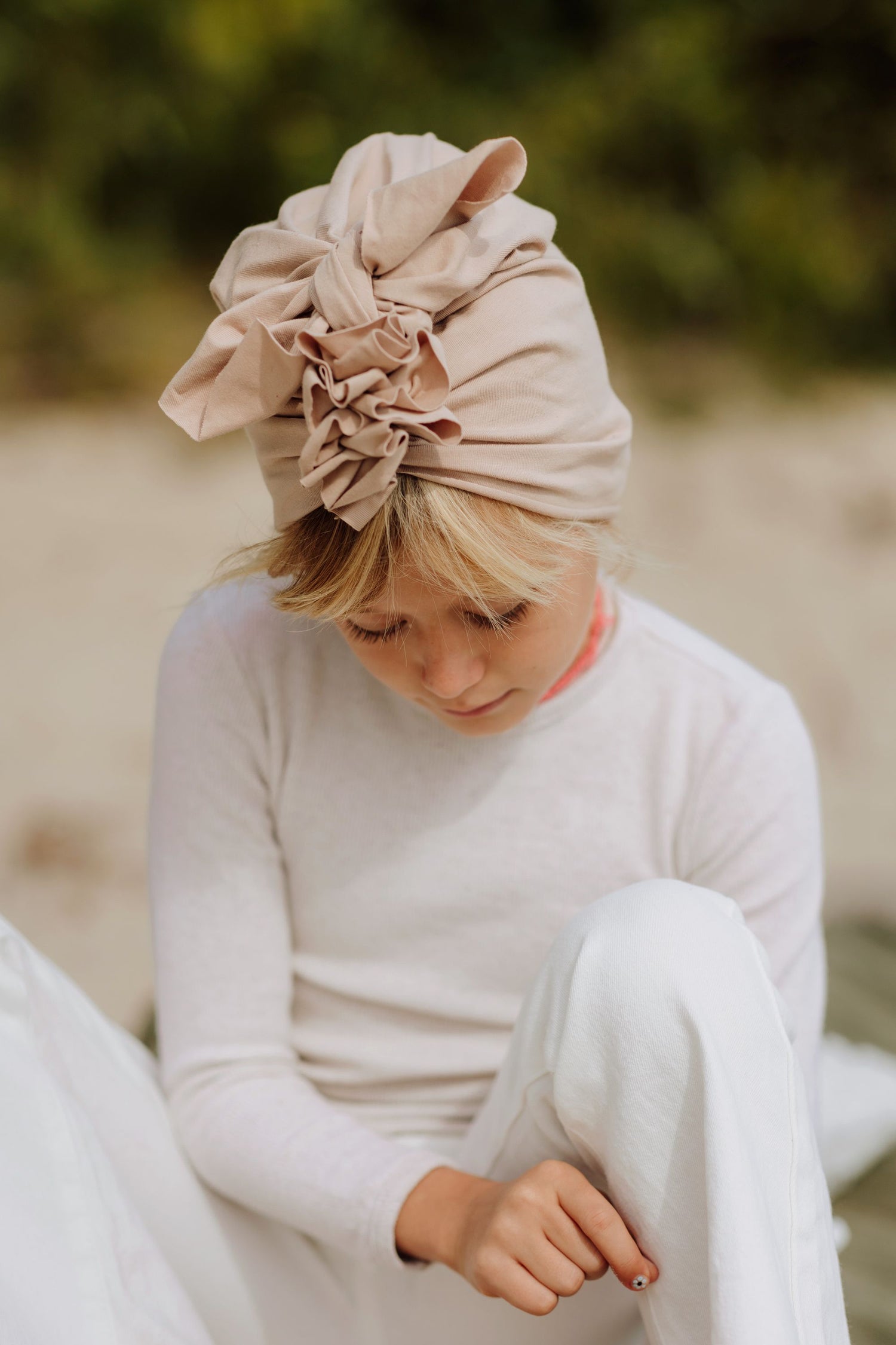 One girl wearing white longsleeve and pants and beige turban looking at her pants sleeve. Forest in the background