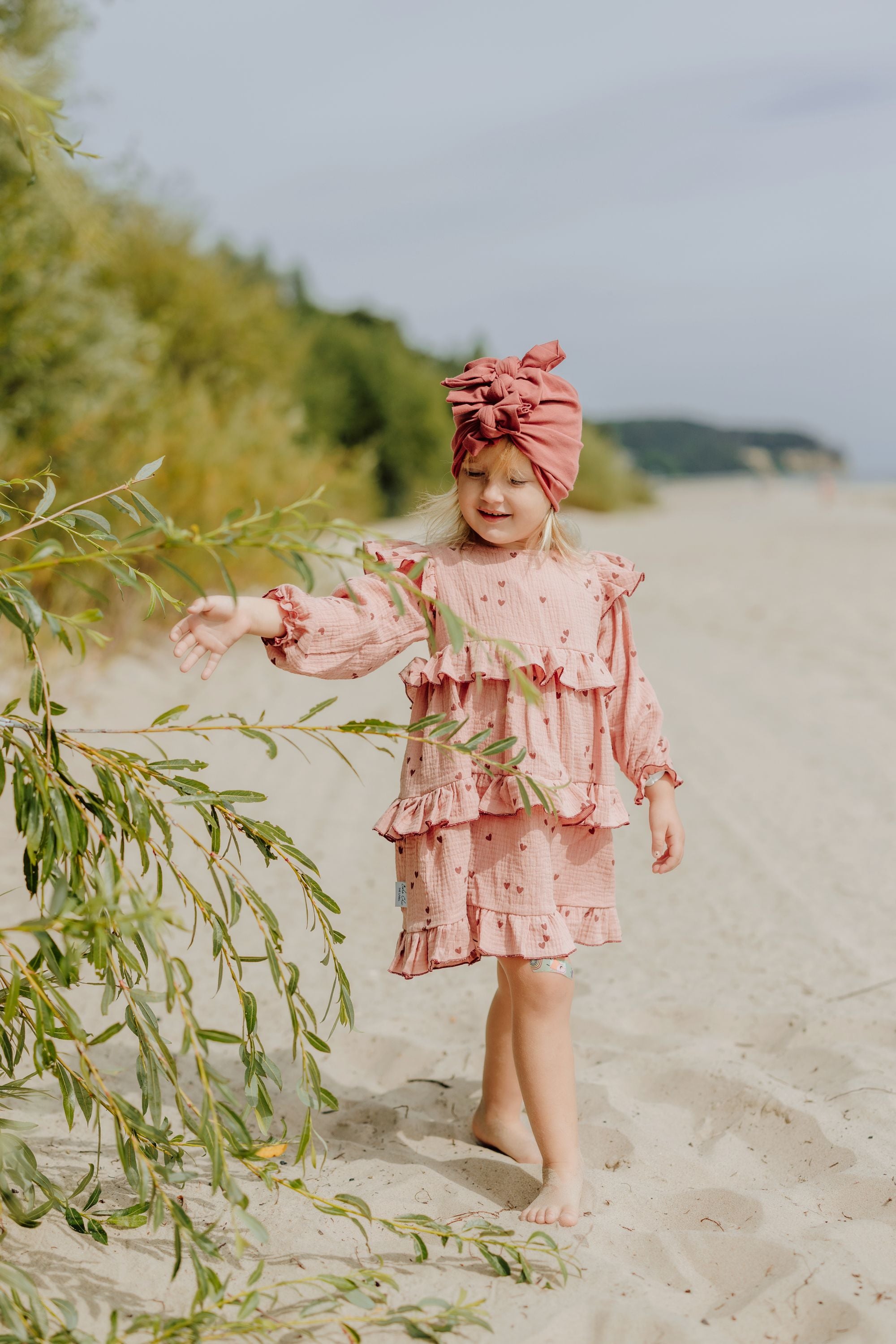 Ein Mädchen ,das auf dem Strand spaziert. Sie hat einen rosa Kleid mit Herzen und eine rote Turbane an. 