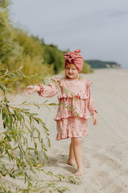 Ein Mädchen ,das auf dem Strand spaziert. Sie hat einen rosa Kleid mit Herzen und eine rote Turbane an. 