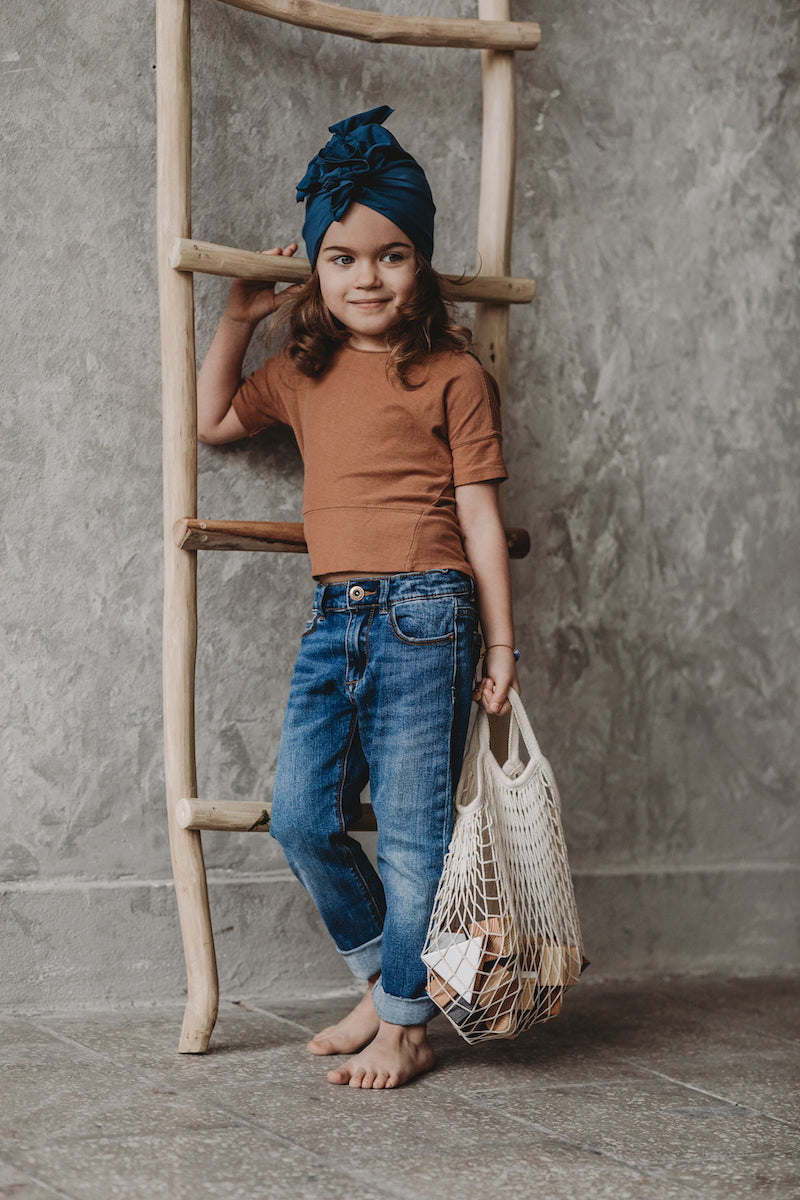 A small girl standing wearing a navy blue turban, jeans and brown tshirt