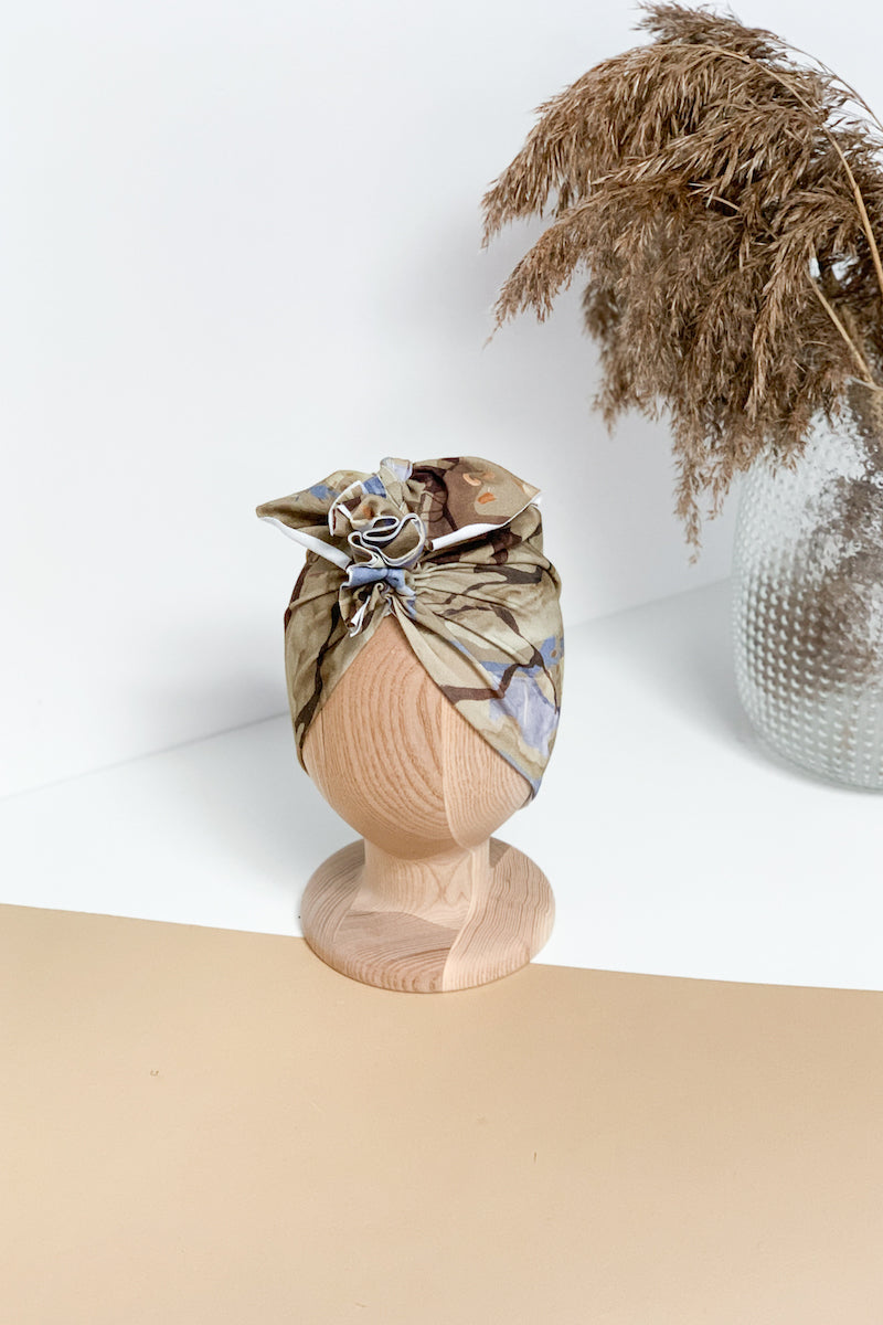 A multicolor turban for children on a wooden mannequin on a white beige background with a silver vase