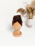 A dark brown velvet turban for children on a wooden mannequin on a white background with white vase