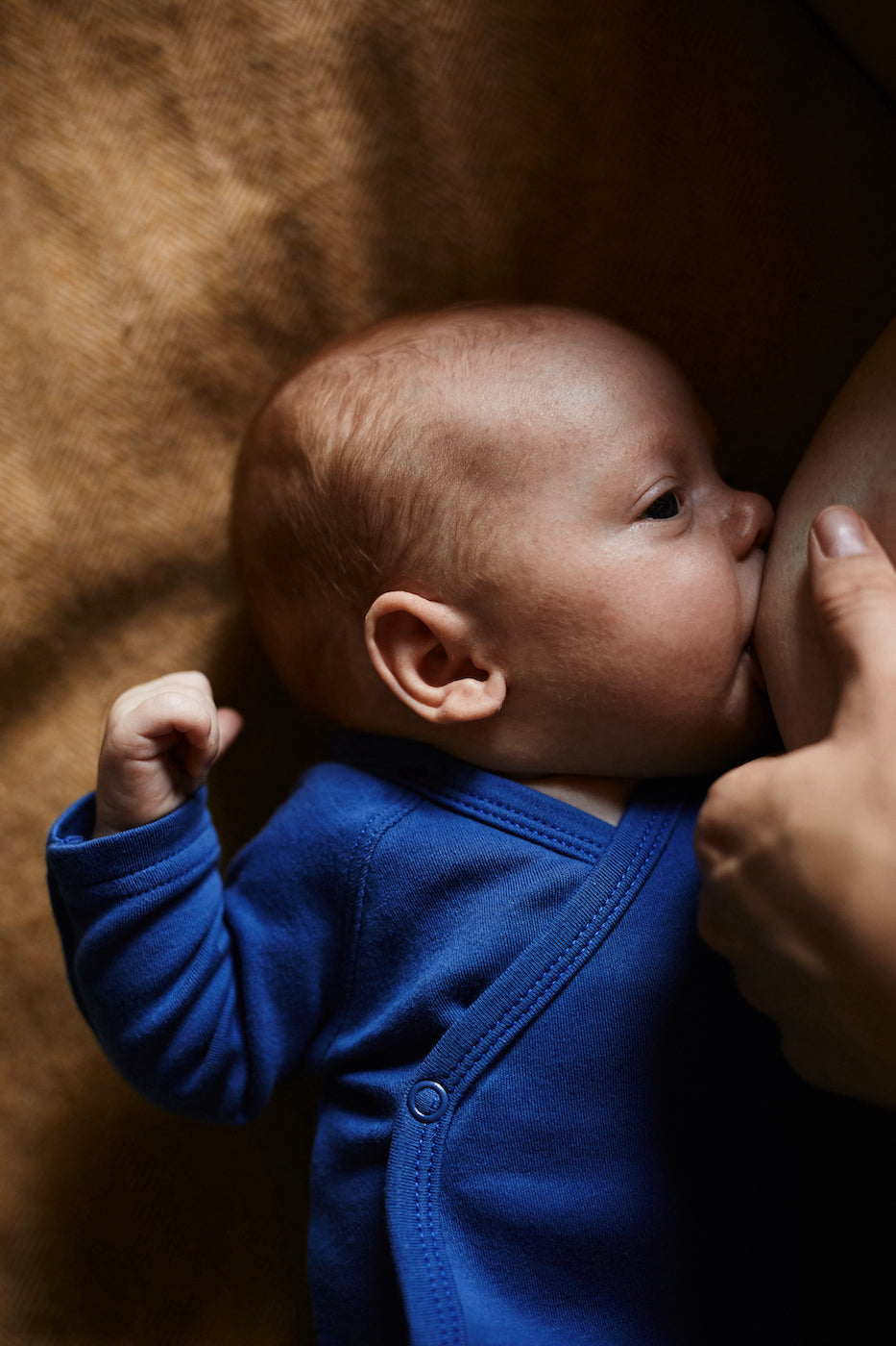 very small baby wearing cobalt blue wrap style bodysuit being breastfed by the mom lying on a brown blanket