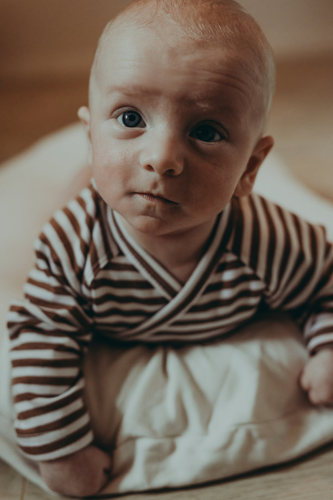 A small baby lying on a belly on a white baby nest wearing a wrap style ecru bodysuit with brown stripes