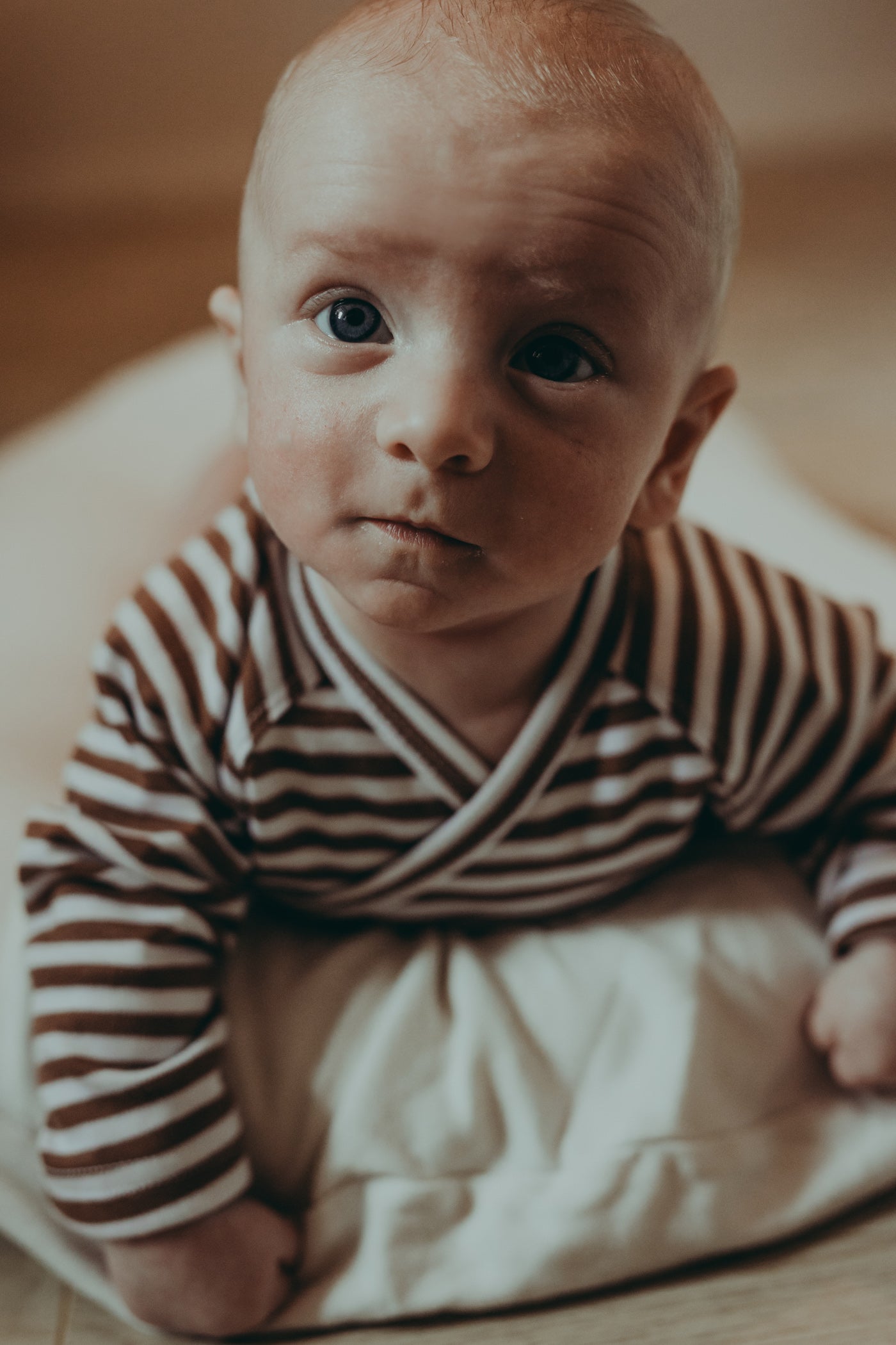 A small baby lying on a belly on a white baby nest wearing a wrap style ecru bodysuit with brown stripes