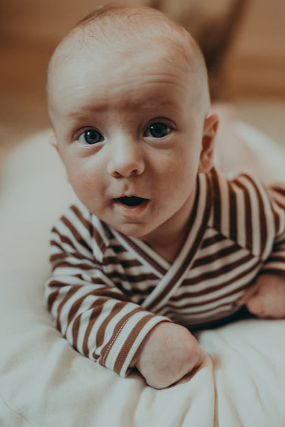 A small baby lying on a belly on a white blanket wearing a wrap style ecru bodysuit with brown stripes