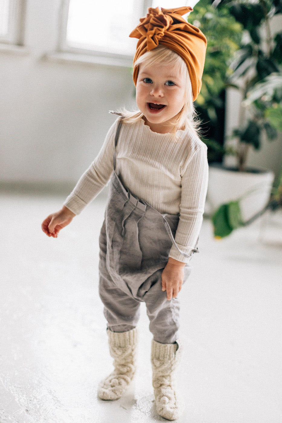 a small blonde girl wearing a beige longsleeve and true orche turban and grey pants, woolen ecru socks