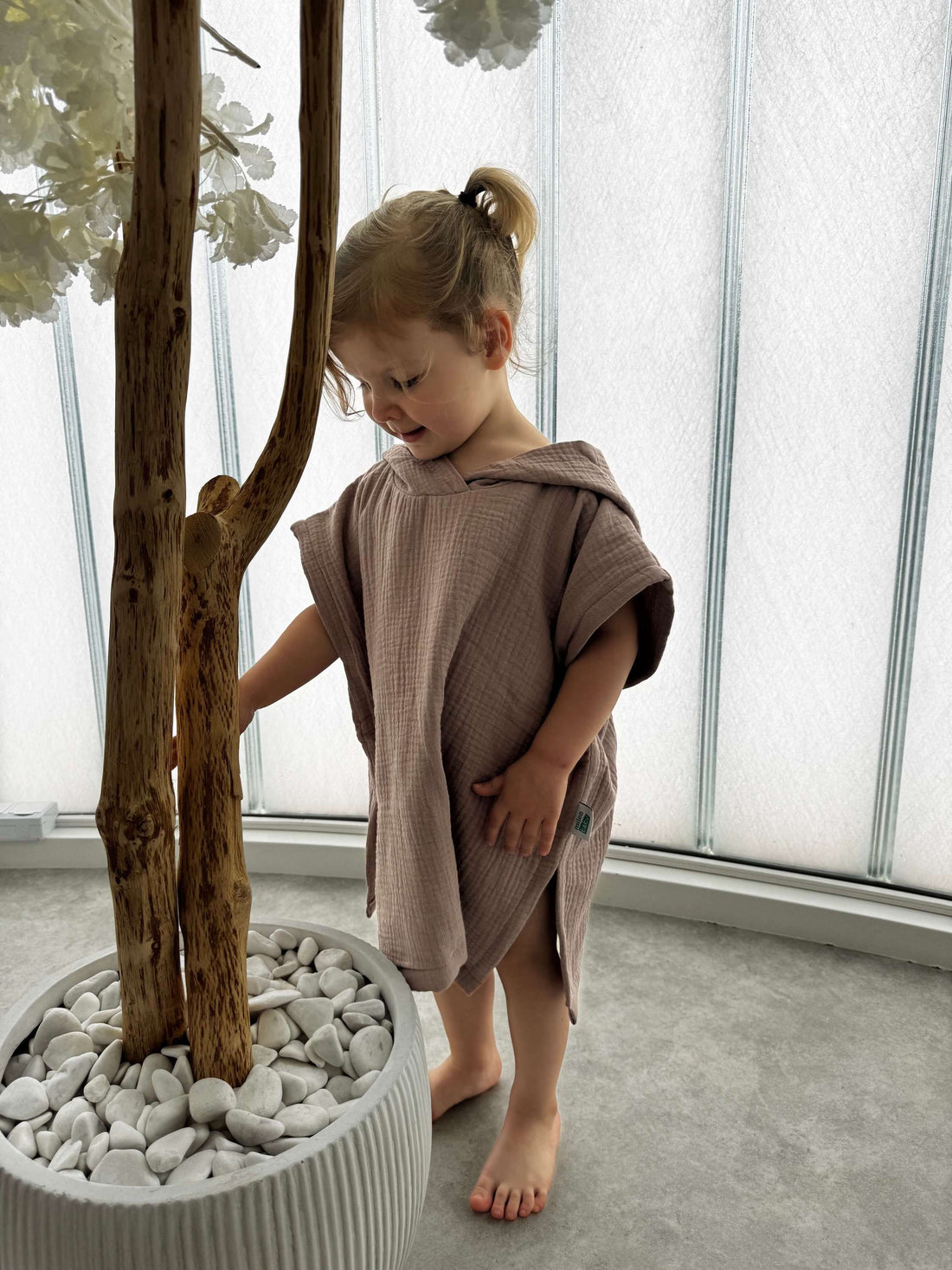Child in a brown towel standing next to a potted plant indoors