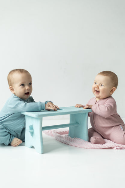 2 babies wearing baby rompers, one in blue and one in pink, smiling and sitting on the blanket