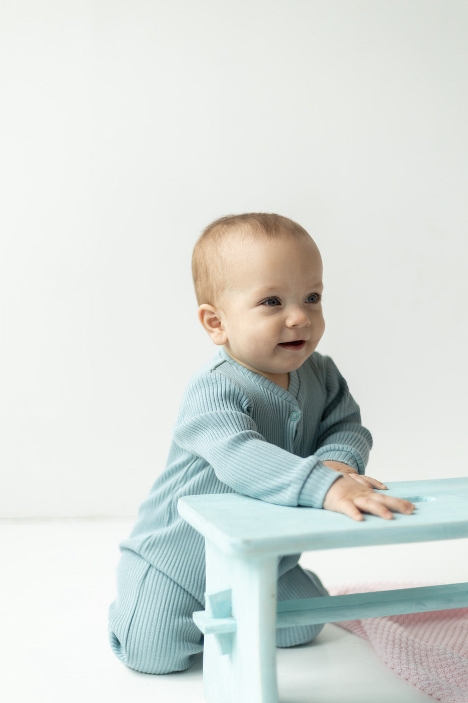 Baby wearing a blue romper climbing on a chair and smiling