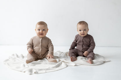 two babies smiling and sitting on the white blanket wearing beige and brown rompers