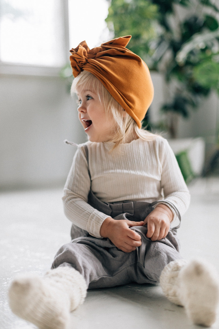 a small blonde girl wearing a beige longsleeve and true orche turban and grey pants, woolen ecru socks is sitting on a the floor and smiling