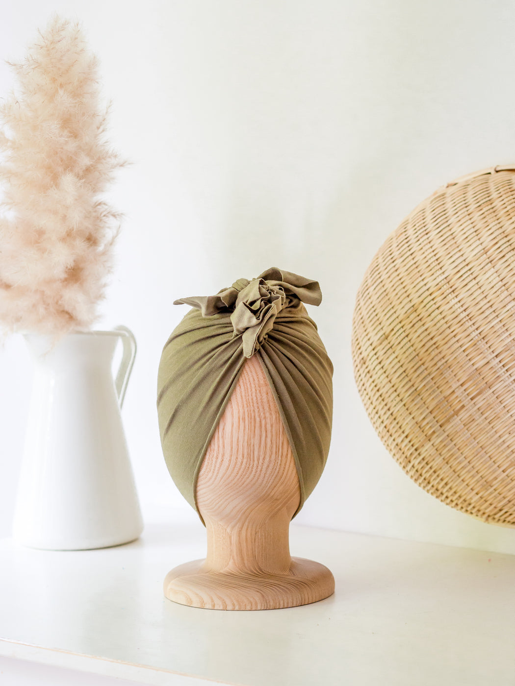 baby turban in color moss green on a wooden head mannequin with a rattan round decoration and white vase in the background