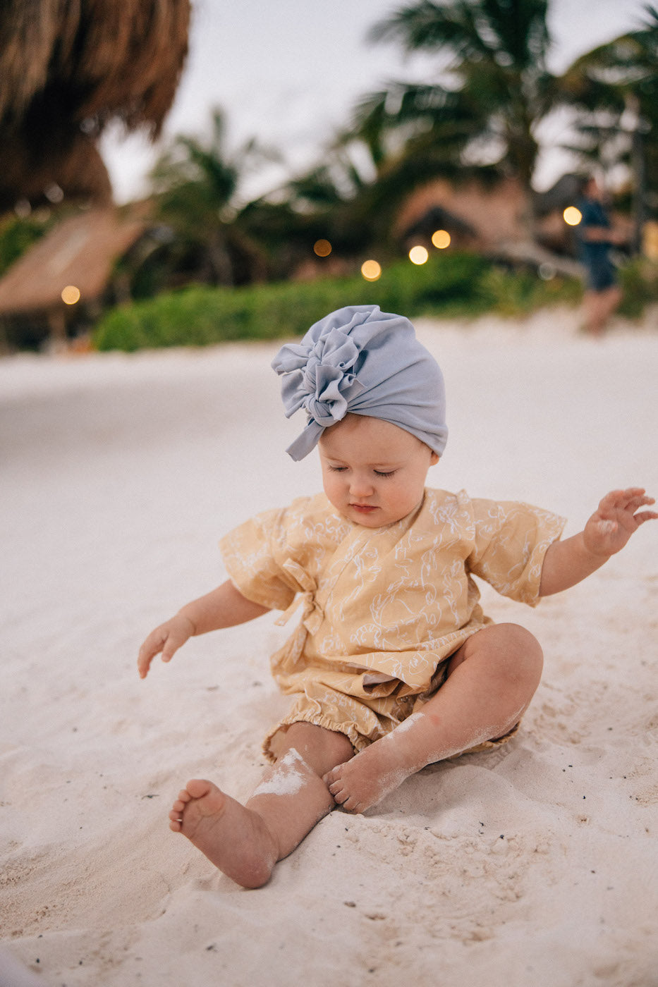 a small baby sitting on the beach wearing a yellow romper and lillac turban