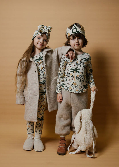 two kids standing on an orange background wearing multicolor clothing and turbans. a boy is holding a bunny backpack and is wearing a crown, girl is wearing a turban and a coat