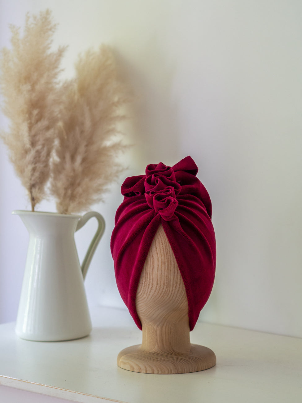 A dark red velvet turban for children on a wooden mannequin on a white background with white vase