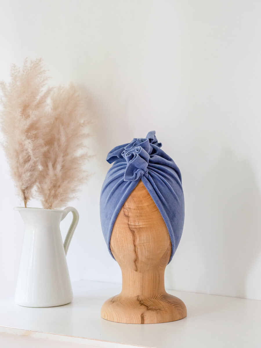 baby turban in color dusty blue on a wooden head mannequin with white vase in the background