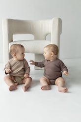 zwei Babys sitzen auf dem Boden vor dem Sessel. Sie habn beige und braue Wickelbody an. 